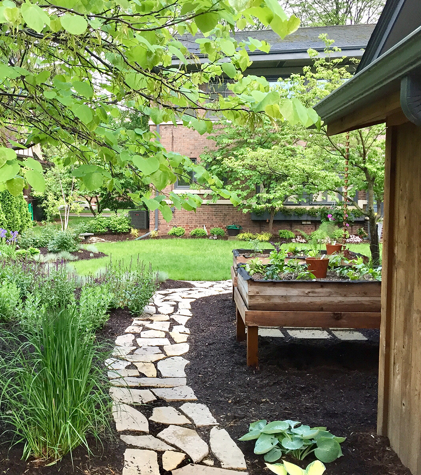 garden-path-1 Serene garden scene with lush greenery, a stone path, and a raised wooden planter. Trees frame a red-brick house in the background.