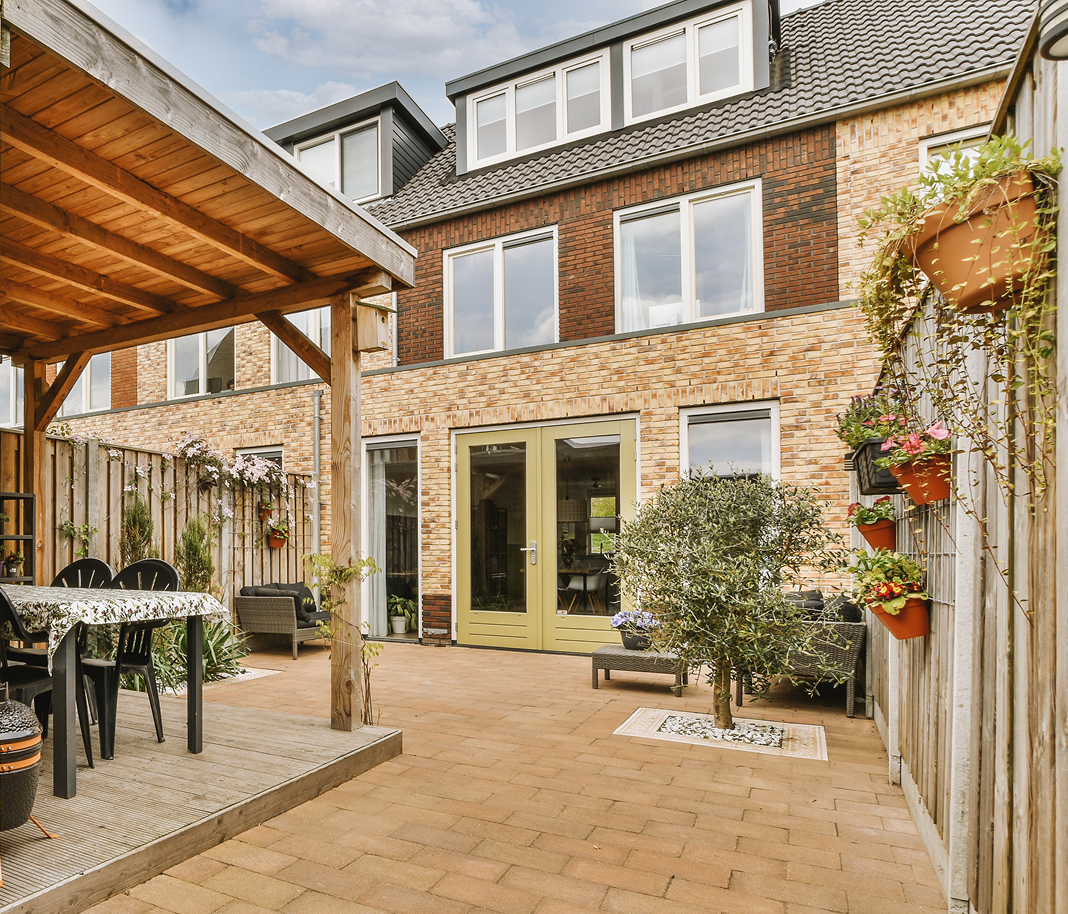 garden-structures Brick townhouse exterior with large windows and a cosy patio. A pergola shelters a dining area with a table and chairs. Potted plants add a vibrant touch.